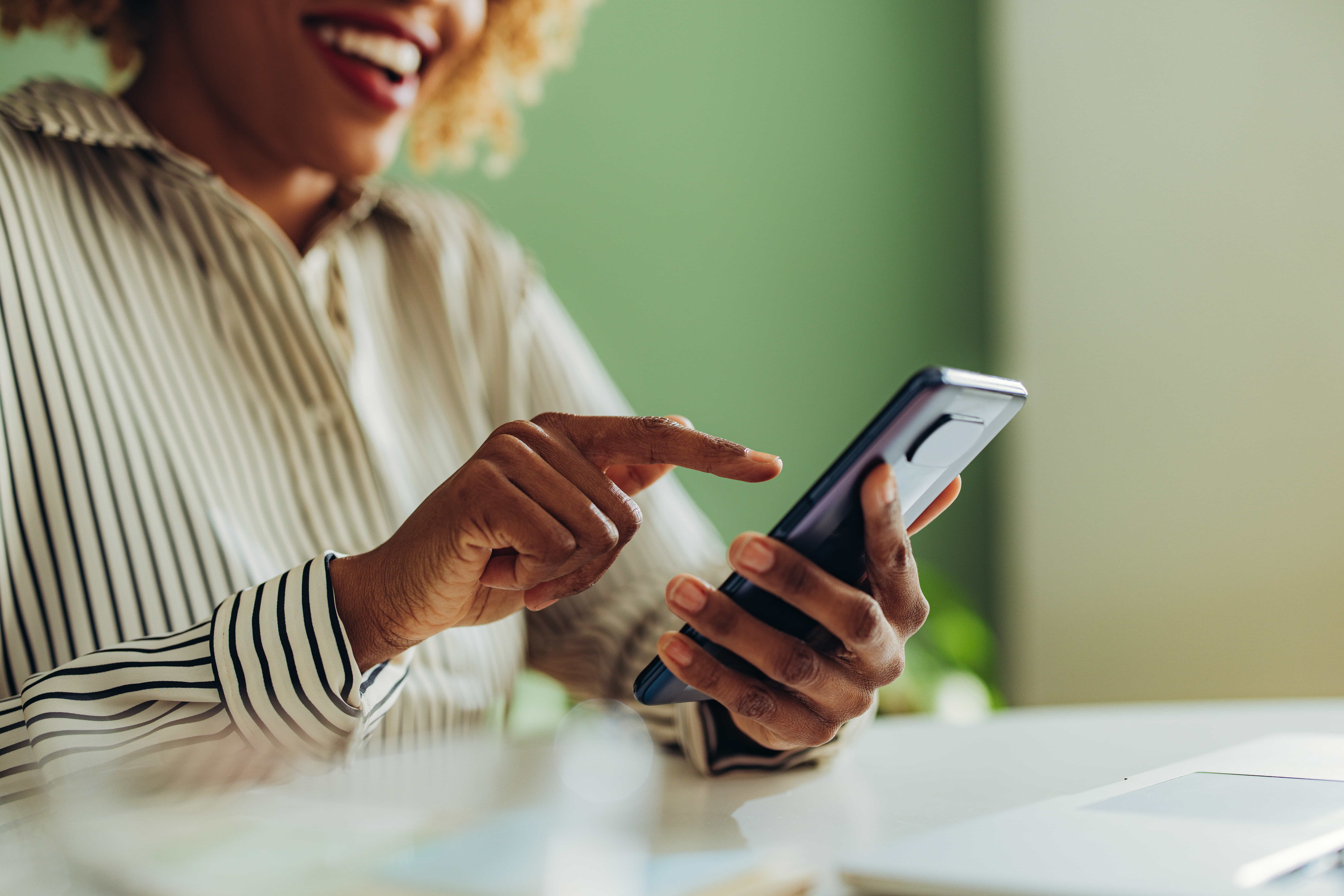 A smiling woman using a smartphone, sitting at a desk with a laptop. She is wearing a striped blouse and tapping the phone screen with one finger, with a blurred green background.